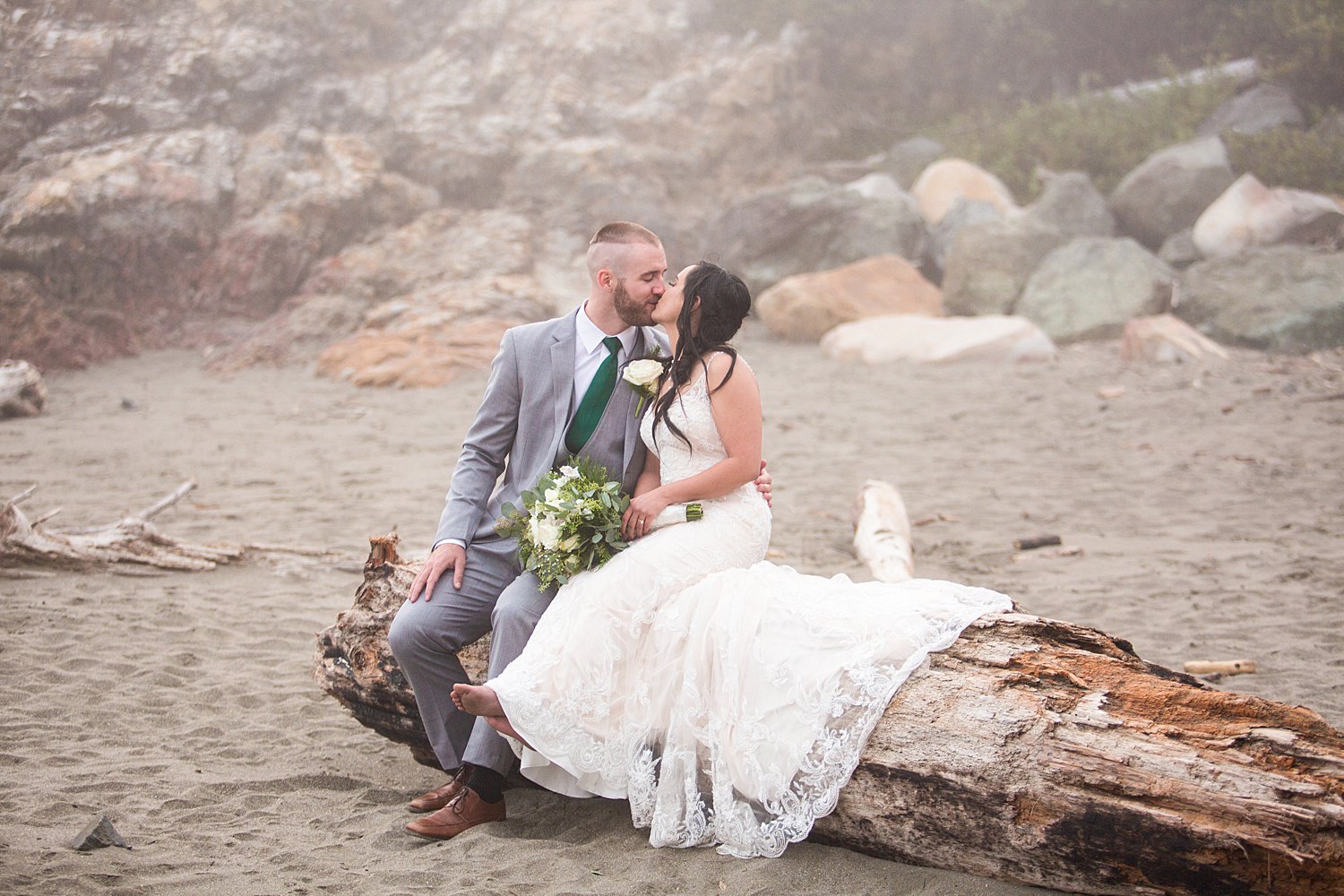 bride and groom kissing sitting on driftwood on the beach